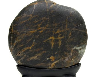 Puede incluir: Una piedra lisa y redondeada de color gris oscuro con vetas de color marrón dorado que recorren toda su superficie. La piedra se muestra en posición vertical, apoyada sobre un soporte de madera negro y curvado con cuatro patas pequeñas.