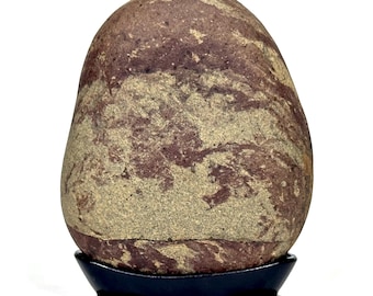 May include: A smooth, oval-shaped stone with a mix of reddish-brown and tan colors, resting on a small, black stand. The stone's surface has a natural, marbled appearance, with swirling patterns and variations in color.