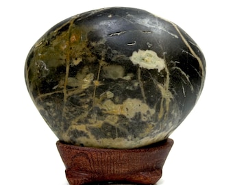 May include: A smooth, rounded dark green and black stone with intricate white veining patterns throughout its surface. The stone is displayed resting securely in a small, carved wooden stand featuring a visible wood grain texture and three feet.