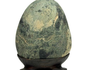May include: An egg-shaped stone sculpture featuring a mottled surface with shades of sage green, dark forest green, and cream-colored veins. The stone is supported by a dark brown, curved wooden stand with three distinct feet at the base.