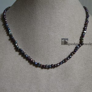Op de afbeelding: Een ketting bestaande uit kleine, onregelmatig gevormde parels. De parels vertonen een mix van donkere kleuren, waaronder zwart, paars en hints van blauw en groen. De ketting is gedrapeerd op een neutraal getinte ondergrond.