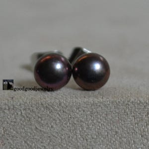 Puede incluir: Un par de pendientes de perlas de color marrón oscuro. Las perlas tienen un brillo lustroso y están engastadas en postes de plata. Los pendientes se exhiben sobre un fondo neutro. La joyería es de goodgoodjewelry.