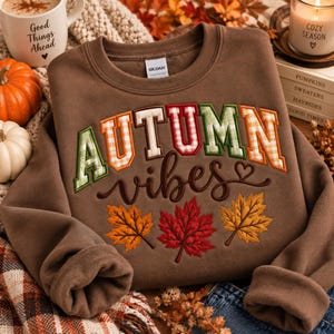 Op de afbeelding: Een bruine sweater met ronde hals voorzien van de tekst AUTUMN in blokletters met groen en oranje ruitpatronen, en het woord vibes in een bruin cursief lettertype met een hartje. Drie geborduurde esdoornbladeren in oranje, rood en geel.