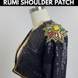 Könnte beinhalten: Schwarze Paillettenjacke mit dekorativem Schulteraufnäher. Der Schulteraufnäher zeigt ein farbenfrohes Blumenmuster mit gelben, rosa, blauen und grünen Akzenten. Die Jacke hat goldfarbene Verzierungen und wird auf einer Schaufensterpuppe präsentiert. Der Text "RUMI SHOULDER PATCH" steht oben.