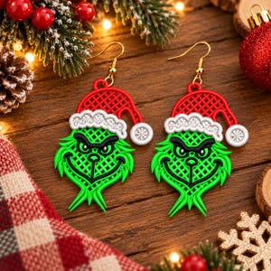 May include: A pair of dangle earrings featuring a green character with a mischievous expression, wearing a red and white Santa hat. The design has a textured, grid-like pattern and is suspended from gold-toned metal earring hooks.