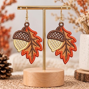 May include: A pair of embroidered dangle earrings featuring a detailed design of an acorn attached to an oak leaf. The acorn has a dark brown cap and a light tan base, while the leaf is rendered in vibrant shades of orange and red.