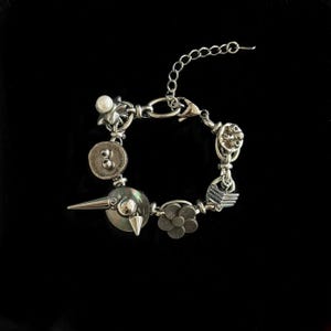 May include: A silver-toned bracelet with various charms, including a star, a button with the number 8, a flower, and a bird-like charm with a spike. The bracelet has a chain extension and a lobster clasp.