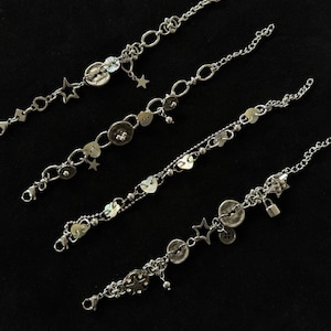 Pode incluir: Quatro pulseiras prateadas com vários berloques, incluindo estrelas, botões e pequenos pingentes. As pulseiras têm diferentes estilos de corrente e fechos. O fundo é preto.