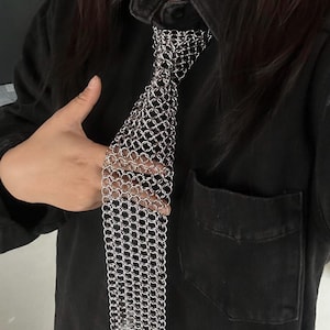 May include: A silver chainmail tie, featuring a woven pattern of small, interlocking metal rings. The tie is displayed against a black denim shirt, showcasing its metallic sheen and unique texture. A modern accessory.