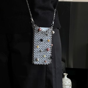 May include: A silver chainmail phone bag with a chain strap. The bag is decorated with colourful beads in shades of pink, black, yellow, and white. The bag is rectangular and has a white interior.