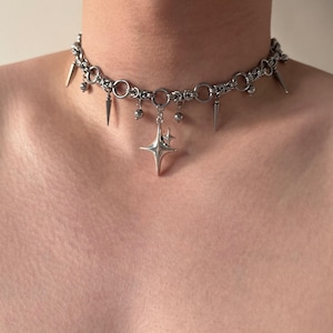 Puede incluir: Collar gargantilla plateado con diseño de cadena, púas, pequeñas cuentas redondas y un colgante de estrella. El collar se lleva alrededor del cuello.