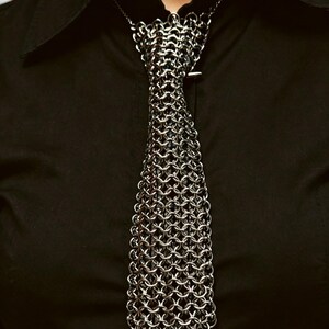 Peut inclure: Une cravate en cotte de mailles argentée, composée de petits anneaux métalliques imbriqués, est mise en valeur sur une chemise noire à col. La cravate présente un aspect texturé et métallique. Un accessoire unique.