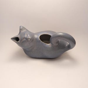 May include: A ceramic teapot shaped like a reclining cat with a speckled blue-grey glaze. The cat's head forms the spout, while its curled tail serves as the handle. An oval opening is located on the back of the cat for filling the vessel.