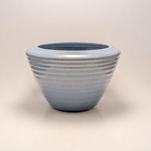 May include: A light blue ceramic bowl with a wide, ribbed design. The bowl has a rounded shape and a smooth, glossy finish. The interior is a darker shade of blue. This decorative bowl is suitable for serving or display.