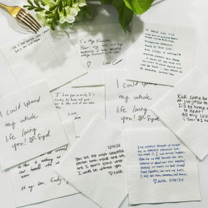 May include: A collection of white paper napkins with handwritten messages in black and blue ink. The napkins feature romantic phrases and dates, with a gold fork and greenery in the background.
