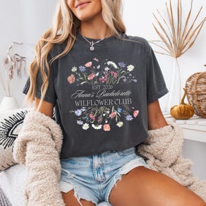 Pode incluir: T-shirt cinza escuro com um design floral e o texto "Anna's Bachelorette WILFLOWER CLUB EST 2026". O design floral inclui flores rosa, roxas e brancas. A camisa &eacute; de gola redonda e manga curta.