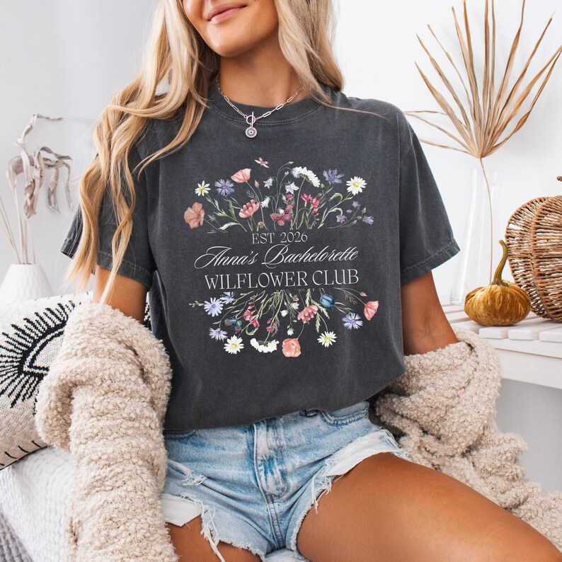 Pode incluir: T-shirt cinza escuro com um design floral e o texto "Anna's Bachelorette WILFLOWER CLUB EST 2026". O design floral inclui flores rosa, roxas e brancas. A camisa &eacute; de gola redonda e manga curta.