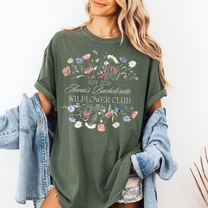Pode incluir: Uma t-shirt verde-oliva com um design floral e o texto "EST 2026 Anna's Bachelorette WILFLOWER CLUB". O design apresenta uma variedade de flores coloridas. A t-shirt &eacute; usada com uma jaqueta jeans.