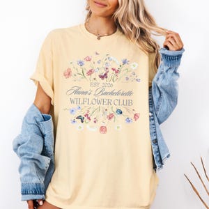 Pode incluir: Uma camiseta amarelo claro com um design floral e o texto "EST 2026 Anna's Bachelorette WILFLOWER CLUB". O design inclui flores rosa, roxas e azuis com borboletas. A camiseta tem mangas enroladas.