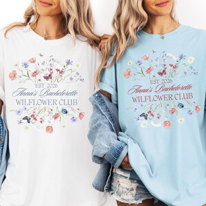 Pode incluir: Duas camisetas, uma branca e outra azul clara, com um design floral e borboletas. O texto nas camisetas diz "EST 2026 Anna's Bachelorette WILFLOWER CLUB". Estas camisetas s&atilde;o para uma despedida de solteira.