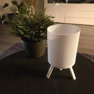 May include: A white cylindrical lamp with a woven texture, supported by three white legs, sits on a dark wooden table. A potted plant with green leaves is in the background, adding a touch of nature to the scene.