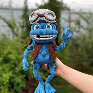 Könnte beinhalten: Eine blaue gehäkelte Charakterpuppe mit brauner Weste, weißer Fliegermütze mit grauer Brille und einer fehlenden Zahnlücke. Die Figur hat dunkelblaue Flecken an den Gliedmaßen, große Augen mit grünen Iris und zeigt ein Friedenszeichen mit der Hand.