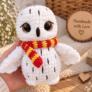 Pode incluir: Um peluche de coruja em croché branco com grandes olhos pretos e um pequeno bico escuro. A coruja apresenta um cachecol às riscas vermelhas e amarelas à volta do pescoço e pequenos detalhes pretos cosidos no corpo, imitando penas na textura.