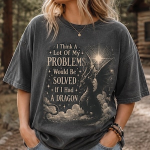 May include: A charcoal gray short-sleeve t-shirt featuring a graphic print of a dragon amidst clouds and stars. The text reads, I Think A Lot Of My PROBLEMS Would Be SOLVED If I Had A DRAGON. The shirt has a crew neckline and a relaxed fit.