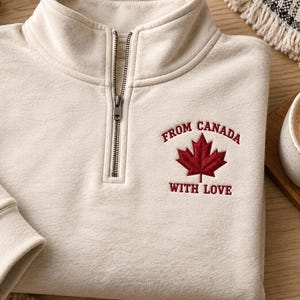 May include: A cream-colored quarter-zip sweatshirt featuring a dark red embroidered maple leaf on the left chest. The text "FROM CANADA" is arched above the leaf, and "WITH LOVE" is embroidered below it in a matching dark red thread.