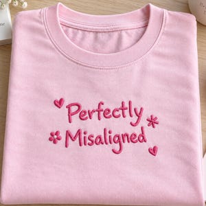 May include: A light pink crew neck t-shirt featuring dark pink embroidery on the chest. The text reads "Perfectly Misaligned" in a cursive font, accented by two small embroidered hearts and two small embroidered flower shapes on either side.
