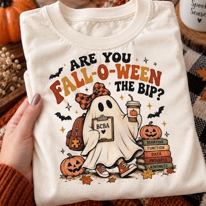 May include: A cream-coloured t-shirt featuring a graphic of a ghost wearing a plaid bow, holding a coffee cup and a clipboard labelled BCBA. The design includes pumpkins, bats, and a stack of books labelled Behaviour, Function, Data, Progress, Kindness.