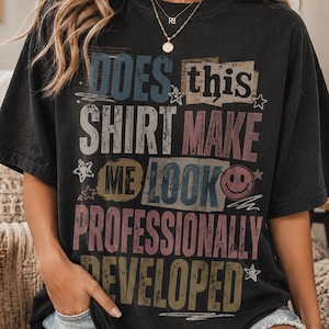 May include: A black short-sleeve t-shirt featuring distressed, multi-colored text that reads: DOES this SHIRT MAKE ME LOOK PROFESSIONALLY DEVELOPED. The design includes a small pink smiley face icon and several star shapes in a vintage-inspired graphic style.
