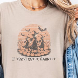 May include: A beige t-shirt featuring a graphic of three witches walking through a pumpkin patch under a sunset sky with bats. The design includes bare trees and the text "IF YOU'VE GOT IT, HAUNT IT" printed in a dark, distressed font below the scene.