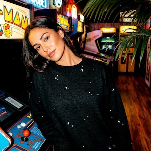 May include: A black crew neck sweater with white star-shaped embellishments. The sweater is worn in an arcade setting, with vintage arcade games and neon signs in the background. The image has a warm, inviting atmosphere.