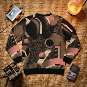 May include: A crew neck sweater with a geometric design in black, brown, pink, and white. The sweater has black cuffs and a black hem. A vintage Polaroid camera, a handheld gaming device, and a cassette player with headphones are also visible.