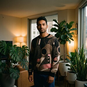 May include: A person wearing a crew neck sweater with a geometric design in shades of brown, black, and pink. The sweater has a black collar and cuffs. The person is standing in a room with plants and natural light.