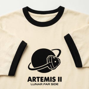May include: Cream-colored ringer t-shirt with black trim on the collar and sleeves. The shirt features a black graphic of a planet with a space helmet and the text "ARTEMIS II LUNAR FAR SIDE" in black.