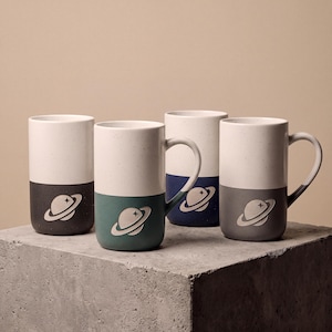 May include: Four tall ceramic mugs with a speckled white upper section and a solid coloured lower section in charcoal, teal, navy, and grey. Each mug features a debossed graphic of a planet with rings on the coloured base and a curved handle.