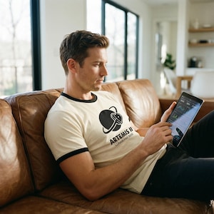 May include: Cream-colored ringer t-shirt with black trim featuring a black graphic of a space helmet and the text "ARTEMIS II LUNAR FAR SIDE". The person is holding a tablet.