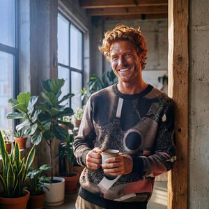 May include: A person wearing a dark gray sweater with a geometric pattern, holding a ceramic mug. The sweater features black, white, and pink shapes. The person is smiling, standing near a window with plants.