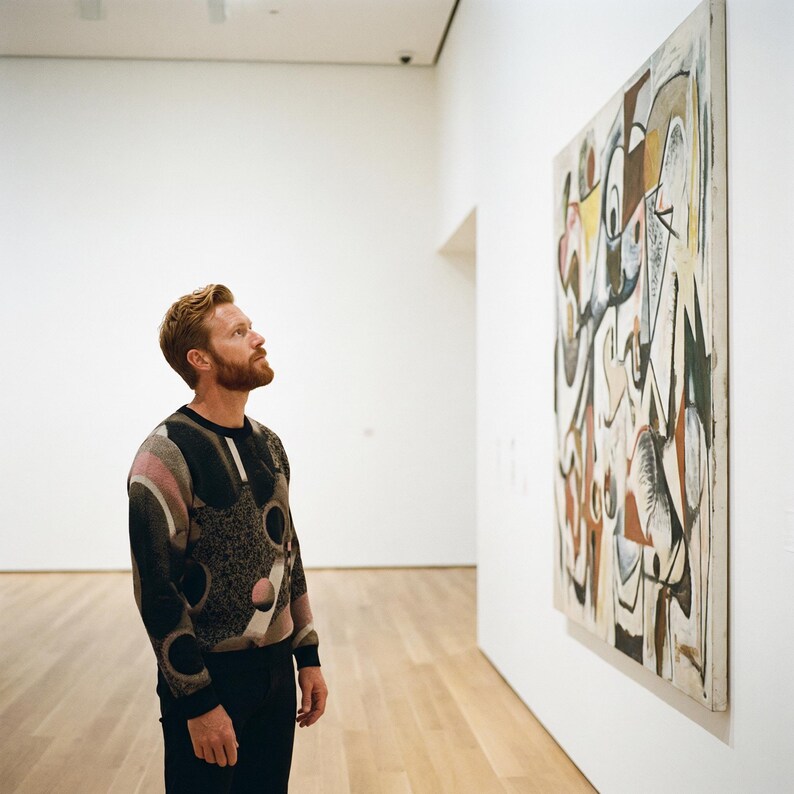 May include: A person in a museum looking at a large abstract painting. The painting features a mix of shapes and colors, including brown, white, yellow, and black. The person is wearing a patterned sweater and black pants.