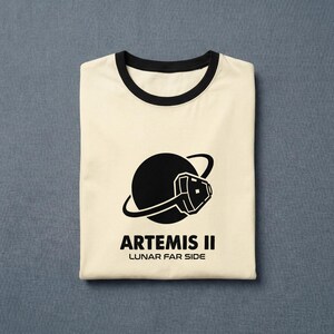 May include: Cream-colored t-shirt with a black collar and cuffs. The shirt features a black graphic of a planet with a stylized space helmet and the text "ARTEMIS II LUNAR FAR SIDE" in black. The shirt is folded neatly.