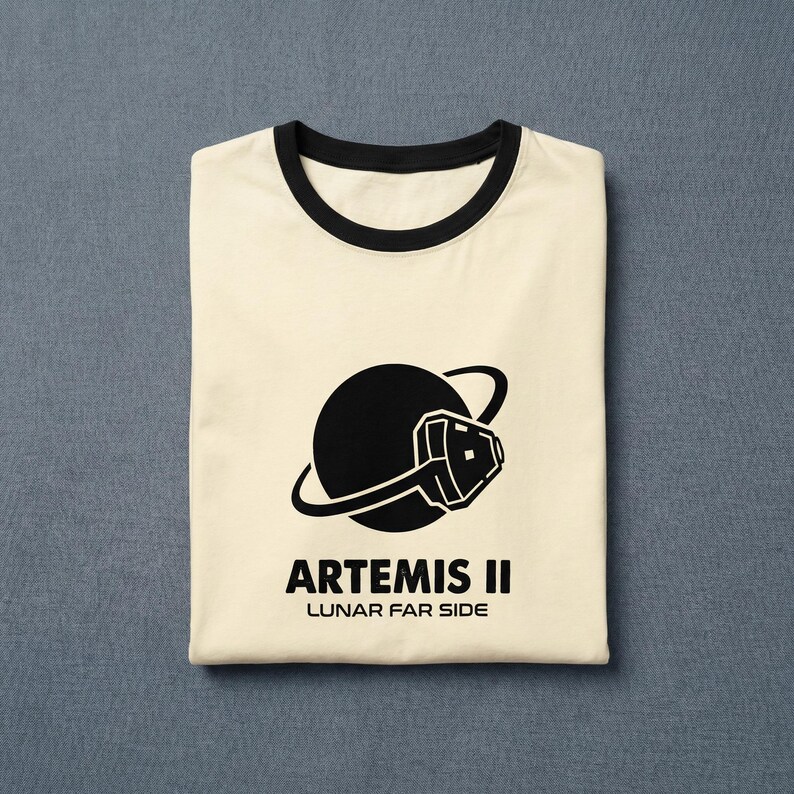 May include: Cream-colored t-shirt with a black collar and cuffs. The shirt features a black graphic of a planet with a stylized space helmet and the text "ARTEMIS II LUNAR FAR SIDE" in black. The shirt is folded neatly.