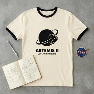 May include: Cream-colored ringer t-shirt with black trim featuring a black graphic of a planet with a spacecraft and the text "ARTEMIS II LUNAR FAR SIDE." A NASA patch and a notebook with diagrams and equations are also visible.