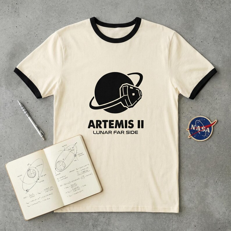 May include: Cream-colored t-shirt with black trim featuring a black graphic of a planet with a space shuttle and the text "ARTEMIS II LUNAR FAR SIDE." A NASA patch and a notebook with diagrams and equations are also visible.