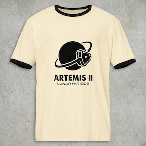 May include: Cream-colored ringer t-shirt with black trim featuring a black graphic of a planet with a stylized spacecraft and the text "ARTEMIS II LUNAR FAR SIDE" in black. The shirt has short sleeves and a crew neck.