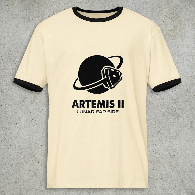 May include: Cream-colored ringer t-shirt with black trim featuring a black graphic of a planet with a stylized spacecraft and the text "ARTEMIS II LUNAR FAR SIDE" in black. The shirt has short sleeves and a crew neck.