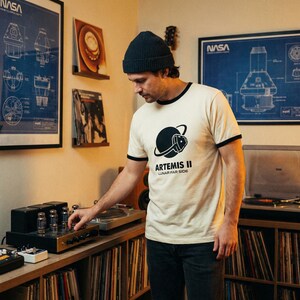 May include: Cream-colored t-shirt with black trim and a graphic of a planet and a space helmet, with the text "ARTEMIS II LUNAR FAR SIDE." The person in the image is wearing a dark beanie and dark jeans. NASA blueprints and vinyl records are in the background.