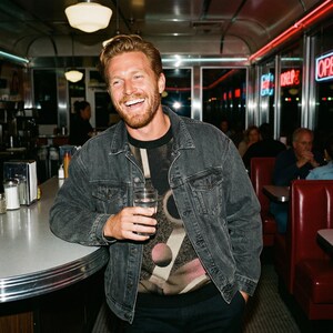 May include: A denim jacket in a dark wash, worn over a graphic print top. The jacket has a classic design with buttoned cuffs and pockets. The person is holding a glass of water in a diner setting.