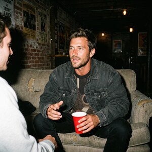 May include: A man wearing a dark denim jacket and black pants, seated on a worn sofa, holding a red cup. The background features brick walls and vintage posters, creating a casual, vintage atmosphere.